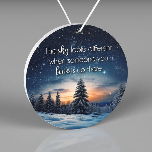 Round Christmas ornament with a night sky, stars, snow and pine trees Words say &quot;The sky looks different when someone you love is up there&quot;
